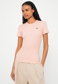 Różowy prążkowany t-shirt z krótkim rękawem, okrągłym dekoltem i zielonym logo na lewej piersi. Tkanina jest miękka o gładkiej fakturze.