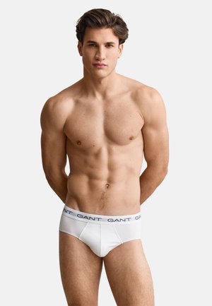 Homme musclé debout avec les bras derrière le dos, portant un slip blanc marqué GANT sur un fond clair uni.