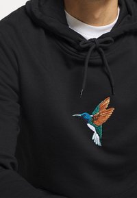 Schwarzer Kapuzenpullover mit Zugkordel, der auf der Brust einen bunten gestickten Kolibri in Blau-, Grün-, Orange- und Weißtönen zeigt.