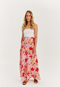 Žena s dlhými blond vlasmi, oblečená v bielom topu bez ramienok, s kvietkovanou maxi sukňou v rúžovej a červenej farbe, hnedými sandálmi a zlatým náhrdelníkom.