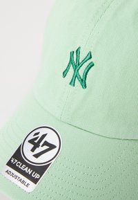 Justerbar mintgrønn baseballcaps laget av stoff; har brodert grønn logo på forsiden og et svart og hvitt merke med detaljer.