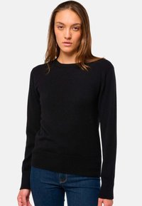 Femme portant un pull noir uni à manches longues et un jean bleu, se tenant devant un fond gris clair uni.