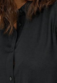 Svart skjorta med krage, som har en slät textur, framknäppning och svarta knappar. Tyget verkar mjukt och lätt.