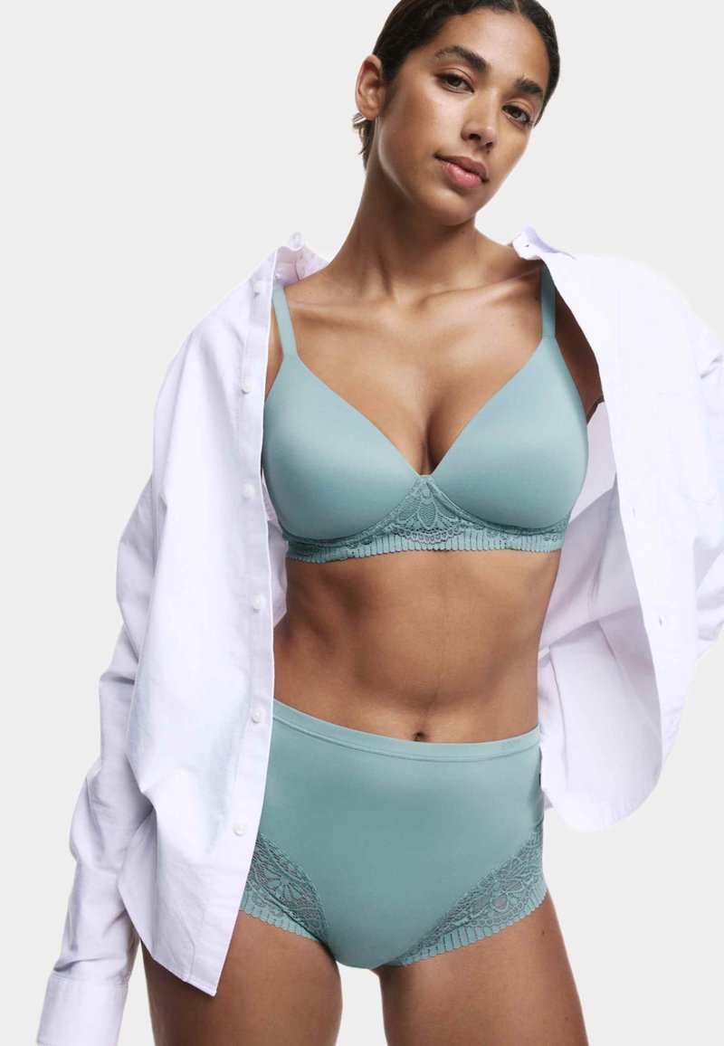 Un reggiseno azzurro chiaro e una slip alta abbinata presentano dettagli in pizzo, una trama liscia e spalline regolabili, indossati sotto una camicia bianca sbottonata.