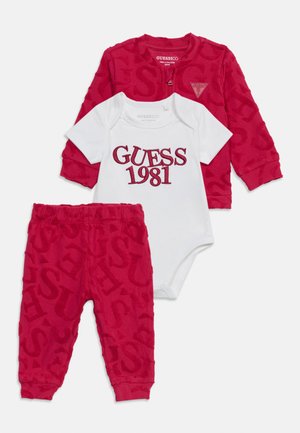 Veste et pantalon bébé rouges avec lettrage en relief « GUESS », associés à un body blanc brodé de « GUESS 1981 » en rouge.