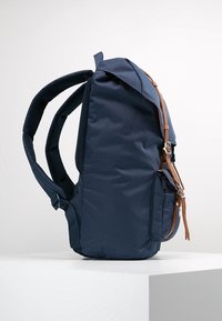 Granatowy plecak z gładką tkaniną, z brązowymi skórzanymi akcentami, regulowanymi wyściełanymi paskami oraz boczną kieszenią na zamek.