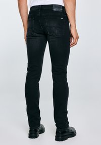 Férfi vékony fekete farmernadrágban és fekete csizmában, kezét lazán oldalt tartva, fehér háttér előtt áll.