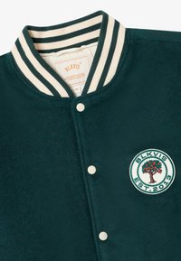 Tume roheline varsity jakk koorevärvi ja roheliste triipudega kraega, koorevärvi nupud ja ümmargune patch puu ning "BLKVIS EST. 2013" tekstiga.