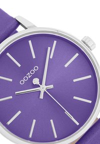 Purple watch with a round dial, silver hour markers, and hands. Features a smooth purple strap and a polished silver case. Brand logo present.