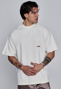 T-shirt en coton blanc surdimensionné à manches courtes, avec un petit logo rouge sur la poitrine et un col rond. Texture lisse et douce.