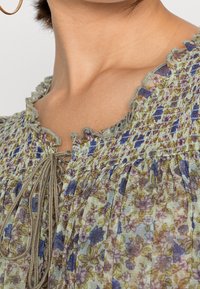 Blommig blus med grönaktig botten, prydd med lila och gula blommor, volangkrage, snörning framtill med dragsko och plisserad textur.