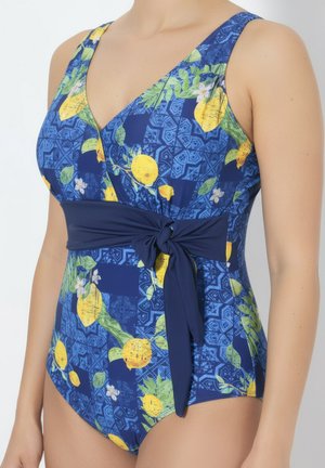 Femme portant un maillot de bain une pièce bleu à motif de citron et de feuilles, avec un profond décolleté en V et une ceinture nouée à la taille.