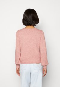 Rosa gestrickter Pullover mit langen Ärmeln, gerippten Bündchen und Saum, der eine weiche Textur aufweist. Getragen mit hellblauen Jeans.