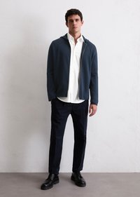 Navy zip-up hoodie, white button-up shirt underneath, black trousers, and black lace-up shoes; smooth textures and casual design.