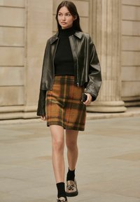 Sort læder cropped jakke over en sort turtleneck, parret med en plaid strikket nederdel i gule, grønne og røde nuancer, samt loafers.