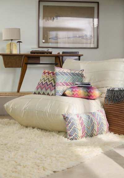 Large cream-colored floor cushion, patterned throw pillows in vibrant colors, a shaggy white rug, and a wooden table with a lamp in the background.