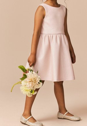 Jeune fille en robe sans manches rose pâle tenant un petit bouquet de fleurs blanches, portant des chaussures Mary Jane scintillantes.