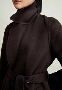 Femme portant un manteau en laine marron foncé ceinturé avec un large col montant, visible du menton jusqu'aux hanches sur fond uni.