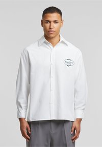 Hvit bomullskjorte med lange ermer og knappelukking foran, med en brodert logo 'Another Cotton Lab' på venstre bryst.