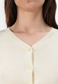Cardigan a coste beige chiaro con scollo a V e tre bottoni. Texture morbida, design aderente e leggero lucido.