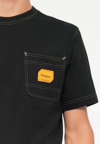 Μαύρο βαμβακερό μπλουζάκι με τσέπη στο στήθος, κίτρινα ραφές και πορτοκαλί ορθογώνιο λογότυπο με την ένδειξη "Replay Inc.".