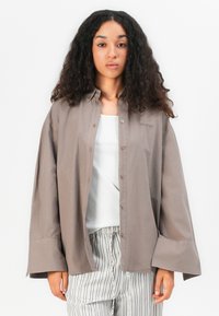 Loose-fitting, long-sleeve shirt in taupe cotton with a button-up front, high cuffs, and subtle branding on the upper left side.