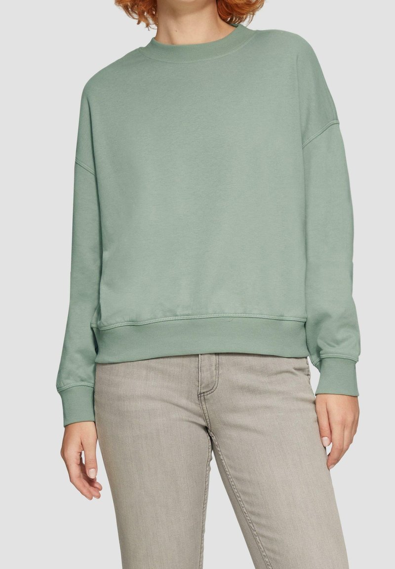Personne portant un sweat-shirt ample vert clair à col rond et un jean gris clair, debout devant un fond uni clair.