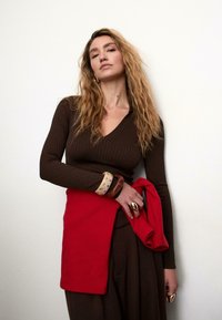 Brązowa, prążkowana bluzka z długim rękawem i dekoltem w serek, zestawiona z luźnymi brązowymi spodniami. Jasnoczerwona tkanina przewieszona przez talię, ozdobiona bransoletkami.