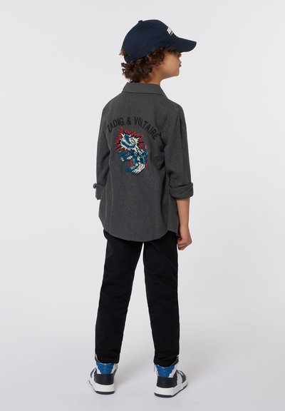 Chemise gris à manches longues avec un design brodé et le nom de la marque au dos, assortie à un pantalon noir et des baskets accentuées de bleu.