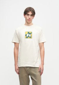 Κρέμα μπλούζα φτιαγμένη από βαμβάκι, με γραφικό σε πράσινο και κίτρινο μέσα σε τετράγωνο. Κοντά μανίκια και άνετη εφαρμογή.