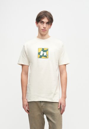 Κρέμα μπλούζα φτιαγμένη από βαμβάκι, με γραφικό σε πράσινο και κίτρινο μέσα σε τετράγωνο. Κοντά μανίκια και άνετη εφαρμογή.