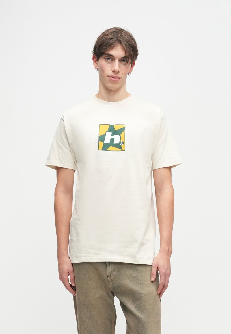 Κρέμα μπλούζα φτιαγμένη από βαμβάκι, με γραφικό σε πράσινο και κίτρινο μέσα σε τετράγωνο. Κοντά μανίκια και άνετη εφαρμογή.