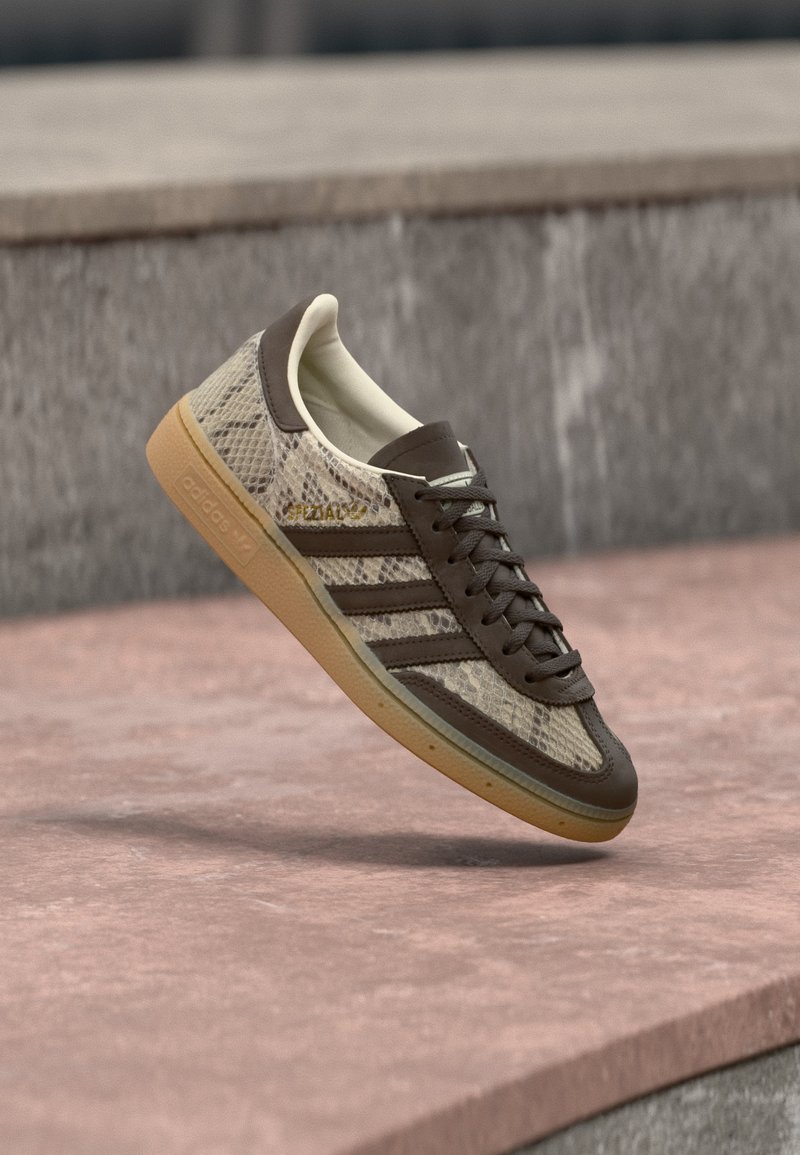 Καφέ και μπεζ αθλητικό παπούτσι Adidas με σχέδιο φιδιού, λαστιχένια σόλα και το κείμενο «SPEZIAL» που αιωρείται πάνω από μια πέτρινη επιφάνεια σε εξωτερικό χώρο.