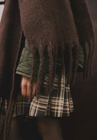 Brown fringed shawl over a green quilted jacket, paired with a plaid skirt featuring dark green and beige stripes.