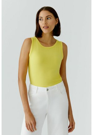 Femme portant un haut jaune sans manches en maille côtelée et un pantalon blanc taille haute, debout devant un fond uni clair.