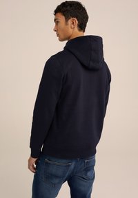 Tamsiai mėlynas džemperis iš medvilnės mišinys, su kengūros kišene, raukšlėmis ant rankogalių ir apatinės dalies. Modelis vilki su šviesiai mėlynomis džinsais.