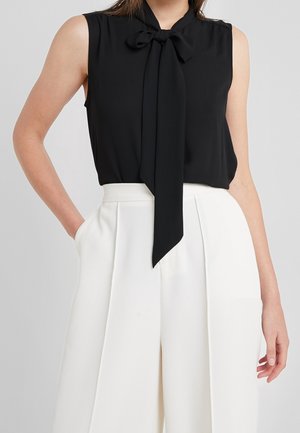 Czarna bluzka bez rękawów z kokardą z przodu, zestawiona z białymi, szerokimi spodniami z wysokim stanem, jedna ręka w kieszeni na jasnym tle.