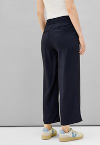 Pantalon large bleu marine avec une finition texturée, taille élastique et deux poches arrière, associé à des baskets beige et bleues.