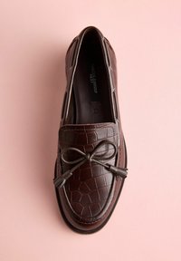 Mocasini maro din piele cu textură de crocodil, prevăzuți cu un accent de fundă și ciucuri. Vârf rotund, plat, pe un fundal roz.