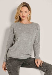 Frau mit blonden Haaren, die einen hellgrauen Pullover mit kleinen weißen Blumenverzierungen und dunkelgrauen Jeans trägt, sitzt vor einem schlichten Hintergrund.
