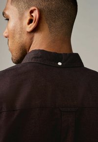 Donkerbruine shirt met een hoge kraag, voorzien van een knoopdetail op de rug en een gestructureerde stof. Het ontwerp is eenvoudig en modern.