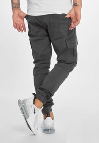 Donkergrijze cargobroek met meerdere zakken, elastische manchetten en een relaxte pasvorm; gecombineerd met wit-zwart sportieve sneakers.