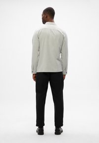 Ανοιχτό γκρι πουκάμισο με γιακά, μακριά μανίκια και κουμπιά στους καρπούς, σε συνδυασμό με μαύρο κομμένα παντελόνια. Το ύφασμα φαίνεται λείο και δομημένο.