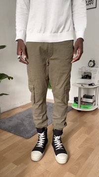 Zielono-oliwkowe spodnie cargo z bocznymi kieszeniami, zwężane nogawki i mankiety u kostek, połączone z czarnymi sneakersami typu high-top z białymi noskami i sznurowadłami.