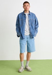 Jeans košulja u plavoj boji, siva majica, svijetloplave šorts hlače i sive patike. Karakteristike uključuju opušteni kroj i ležeran stil.