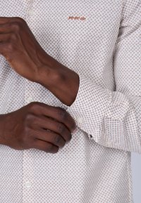 Chemise blanche avec un petit motif paisley marron répétitif ; comprend des poignets à bouton et un petit logo brodé marron sur la poitrine.