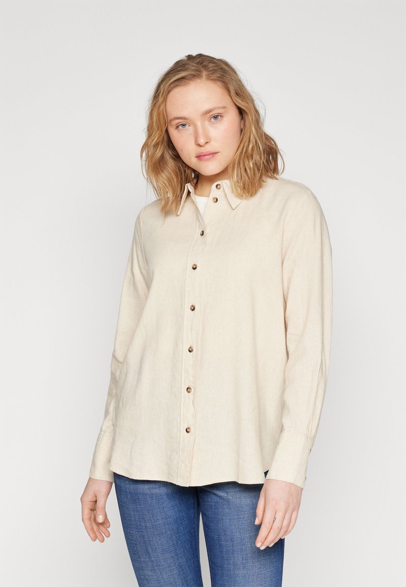 Beige linen button-up shirt with a collared neckline, featuring brown buttons down the front and long sleeves, paired with blue jeans.