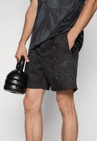Black athletic shorts with a textured floral pattern, featuring side pockets and an adjustable waistband, held by a person beside a kettlebell.