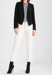 Mujer vistiendo un blazer negro, una blusa gris, unos vaqueros blancos cortos con dobladillos deshilachados y botas de tacón alto negras hasta el tobillo, de pie sobre un fondo blanco.