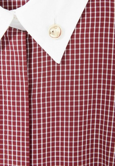 Primo piano di una camicia a quadri borgogna e bianca, con colletto classico e un bottone dorato con logo. Tessuto texture visibile.